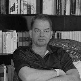 Black and white picture of Chris Adami with his arms crossed looking at the camera. He is sitting in front of a bookshelf full of books. 